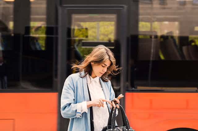 A woman standing beside a bus uses a smart phone while holding a purse