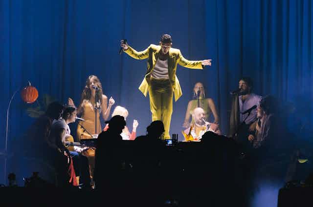 C. Tangana con traje amarillo está sobre una mesa que rodean todos los invitados a su concierto.