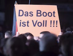 A person holds up a sign that reads the boat is full in German.