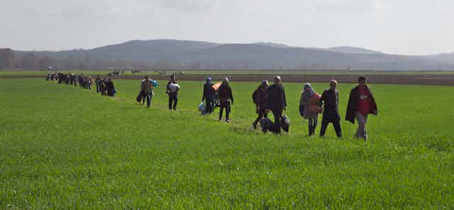 A line of people carrying their belongings walk in a grassy field with hills behind them.