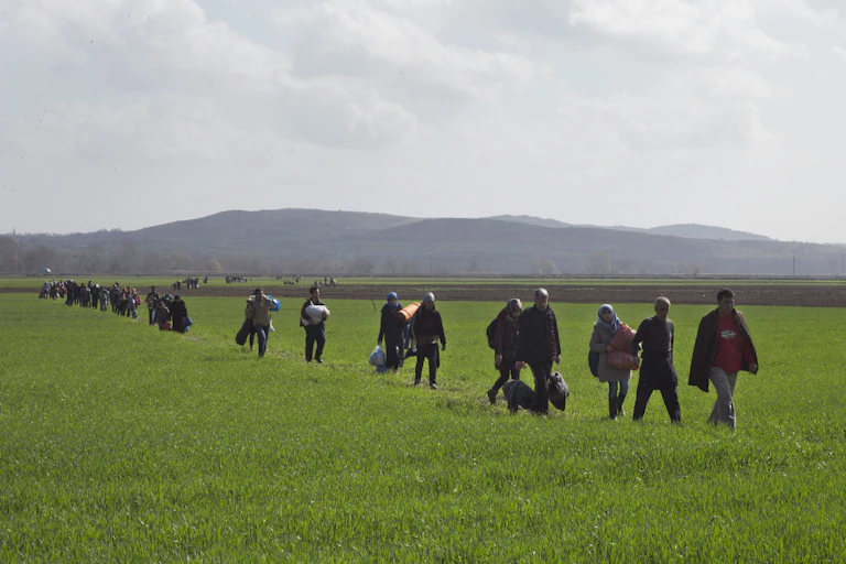 A line of people carrying their belongings walk in a grassy field with hills behind them.