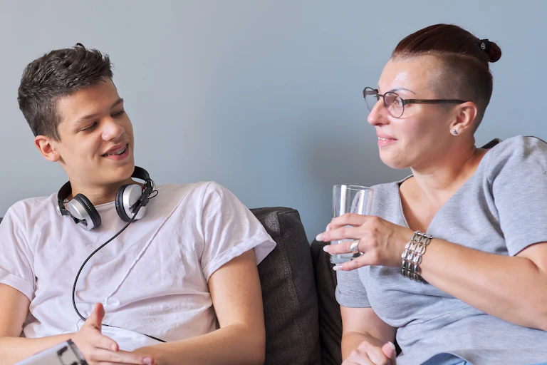 A teen in headphones sits talking to his mom who watches him with a smile on her face.