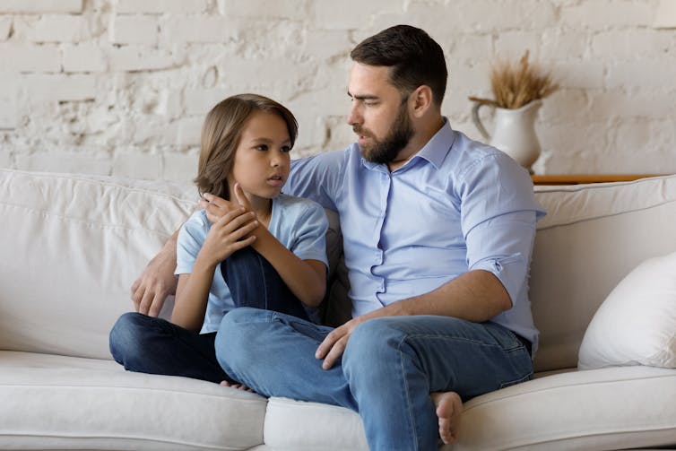 A man with a beard with his arm around a boy sitting on a white sofa