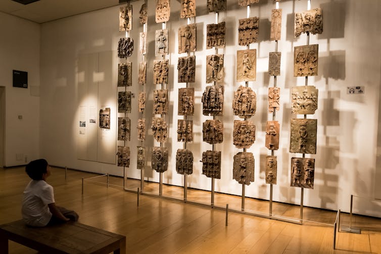 A child sits on a bench infront of a museum display that shows small bronze figures.