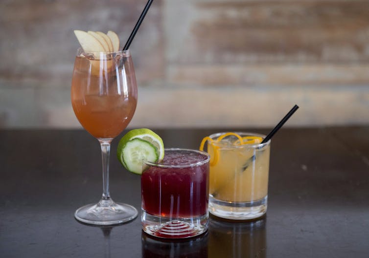 Three cocktails with garnishes: an orange beverage in a wine glass, a red beverage and a yellow beverage in short glasses