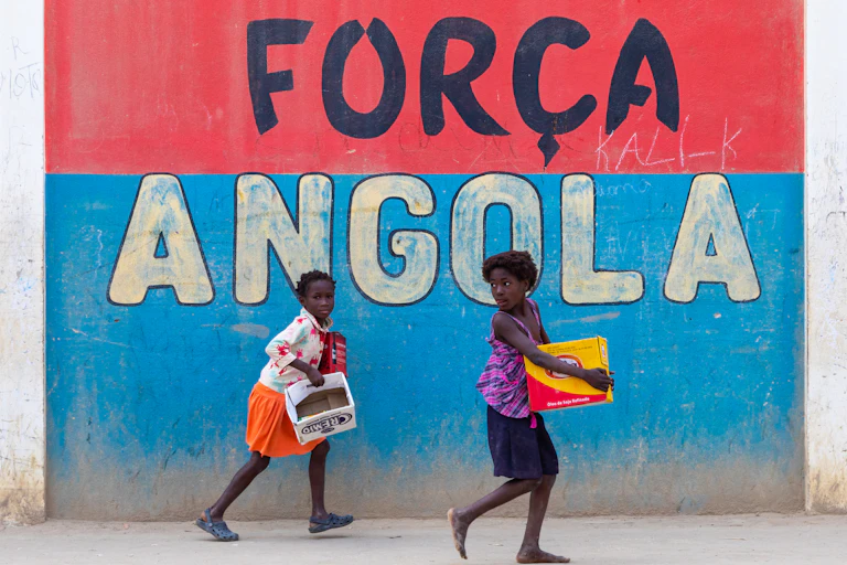 Angolan girls in front of a patriotic slogan on a wall.