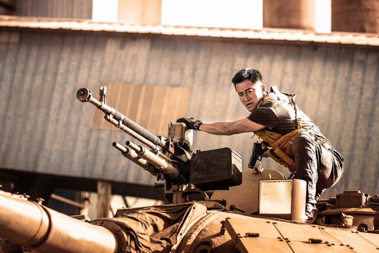 A Chinese man in combat gear crouches on top of an armed vehicle, hand ready on a gun.