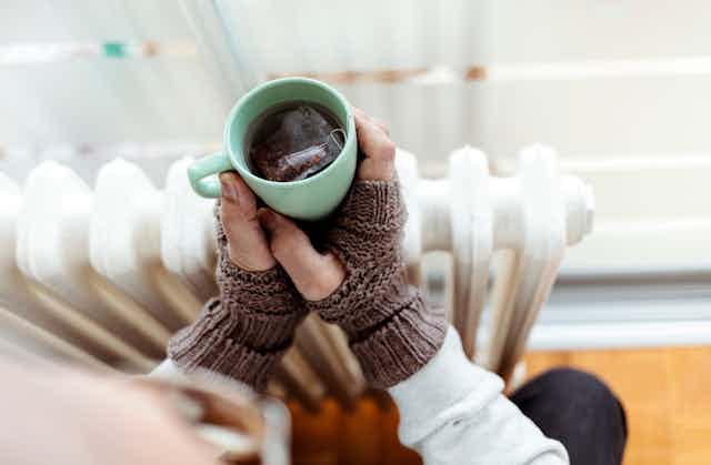 Hands with gloves holding mug of tea over a radiator.