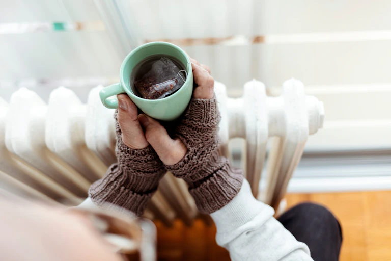 Hands with gloves holding mug of tea over a radiator.