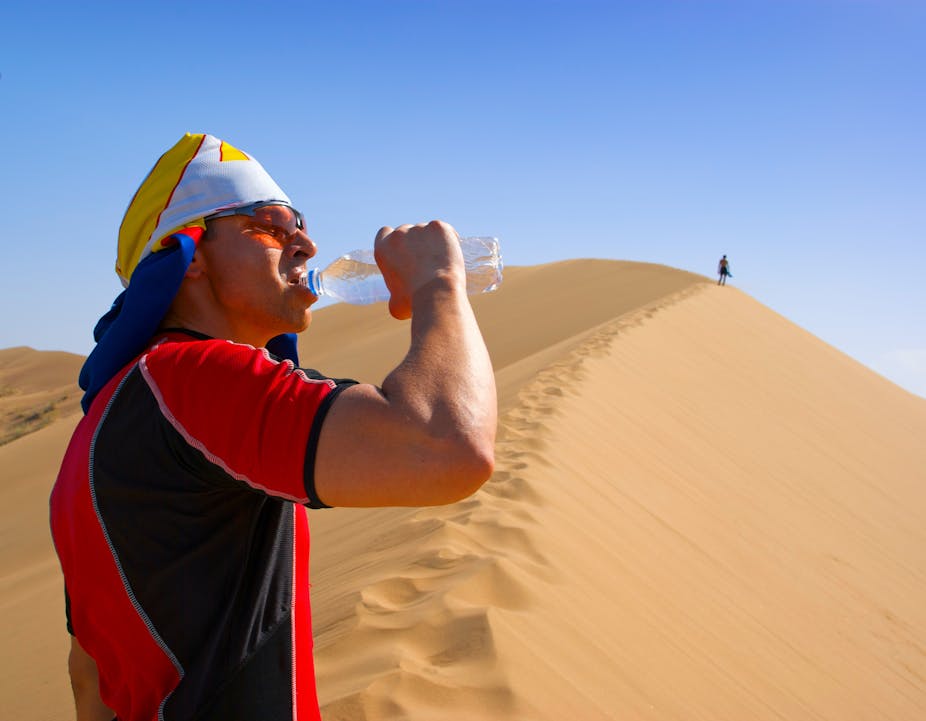 A man standing on top of a desert sand dune drinking from a bottle of water.