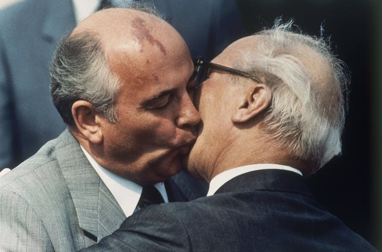 A bald man and another in glasses kiss on the mouth.