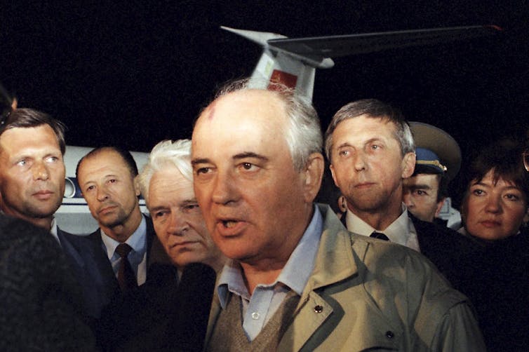A bald man is seen surrounded by other men with a portion of an airplane seen behind them.