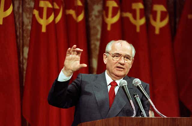 A balding man with glasses gestures as he speaks into a microphone, a row of hammer-and-sickle red-and-gold flags behind him.