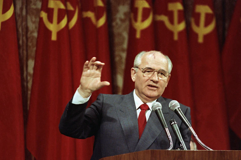 A balding man with glasses gestures as he speaks into a microphone, a row of hammer-and-sickle red-and-gold flags behind him.