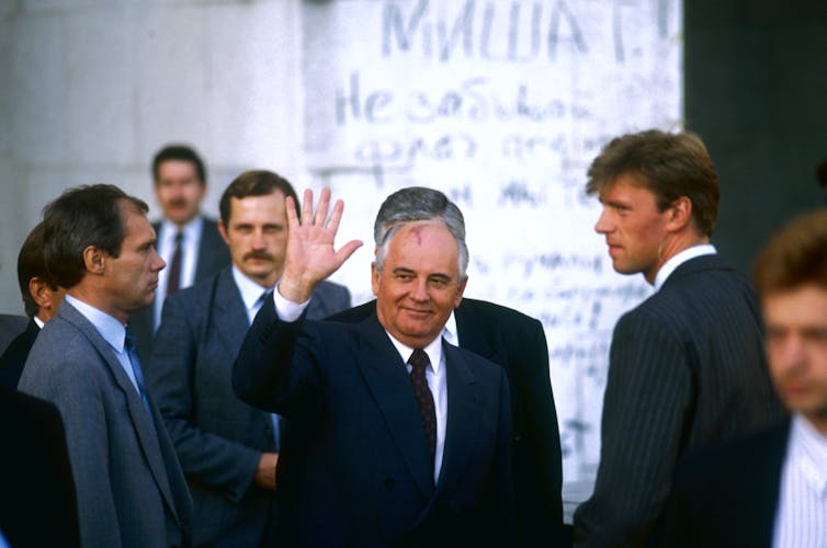 A bald older man with a dark suit is seen waving, with other men around him.