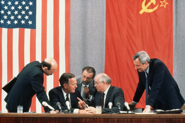 Five older men are seen leaning in toward each other at a table with microphones, sitting in front of Soviet and U.S. flags.