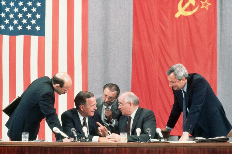 Five older men are seen leaning in towards each other at a table with microphones, sitting in front of Soviet and U.S.A. flags.