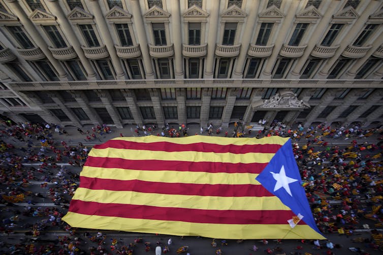 Hundreds of people carry a Catalan red, yellow and blue flag, photographed from above on a city street.