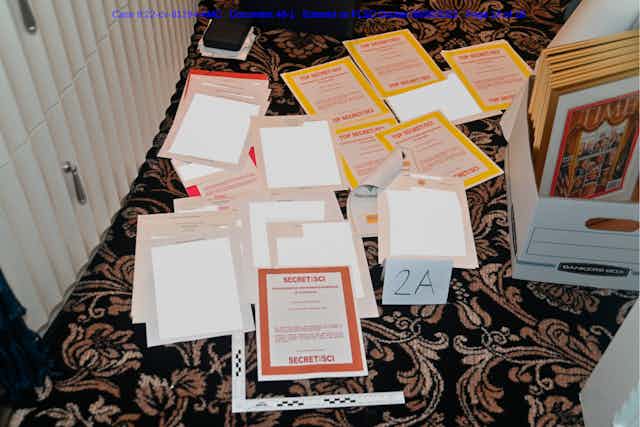 A bunch of papers stacked, with terms like Secret/SCI are shown on a carpeted floor, next to a box containing framed documents, including a TIME Magazine cover. 