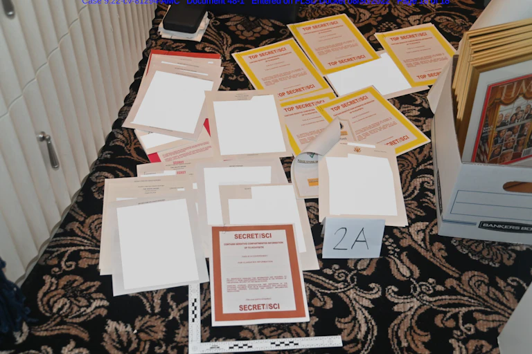 A bunch of papers stacked, with terms like Secret/SCI are shown on a carpeted floor, next to a box containing framed documents, including a TIME Magazine cover.