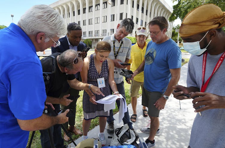 A group of people gather outside a government building and look at a blacked-out document. Some of them hold cameras.