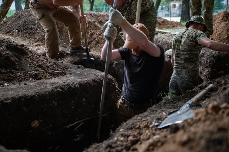 Several men dressed in military gear are using shovels to dig combat trenches in Ukraine.