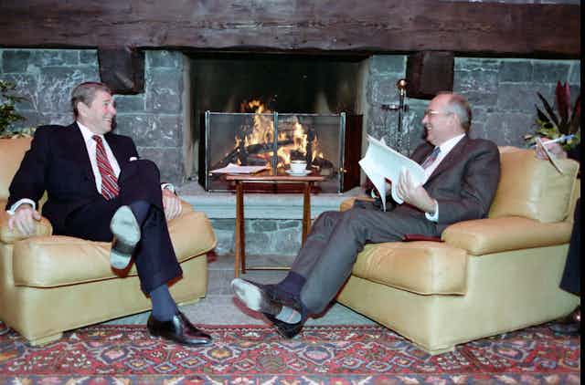 Ronald Reagan and Mikhail Gorbachev sit smiling in armchairs in front of a fire.