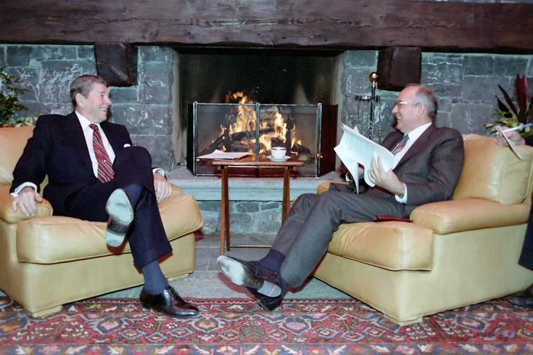Ronald Reagan and Mikhail Gorbachev sit smiling in armchairs in front of a fire.