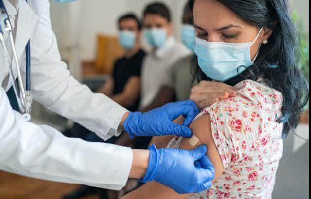 A doctor in white lab coat and blue gloves places Band-Aid on a woman wearing a mask who has just received a COVID-19 vaccine.