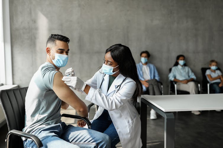 A young man receives a vaccination while other people wait in the background.