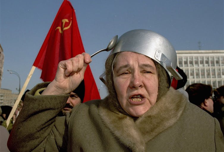 An aging woman with a pot on her head strikes it with a spoon.