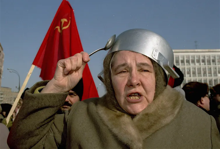 An aging woman with a pot on her head strikes it with a spoon.