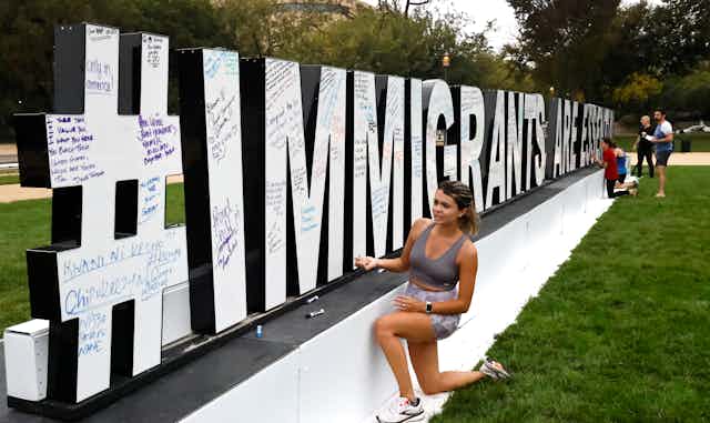 A womankneels next to an outdoor sculpture spelling #immigrants are essential