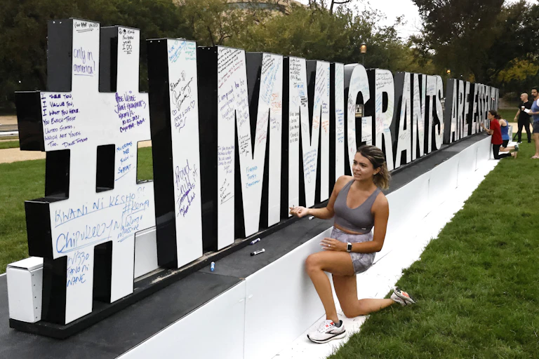 A womankneels next to an outdoor sculpture spelling #immigrants are essential