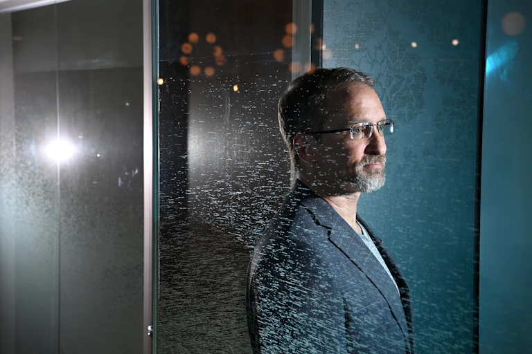 a man with short hair, glasses and a beard wearing a suit jacket seen reflected in a scratched or dirty mirror
