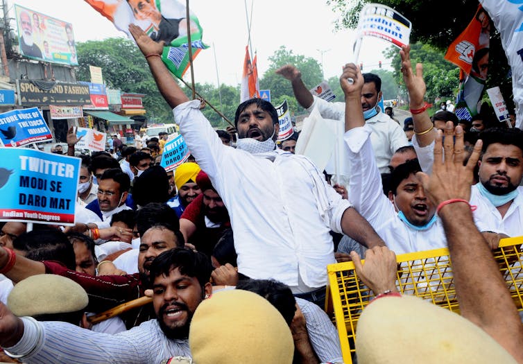 a line of men wearing beige berets in the foreground holds back a crowd of young men shouting and waving banners