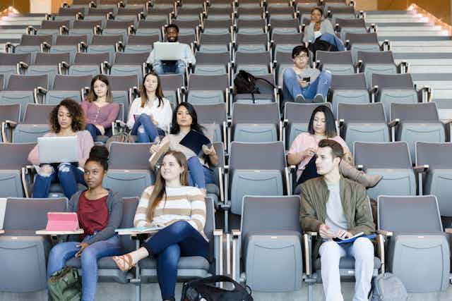 Eleven young college students – mostly women – sit in grey seats in a lecture hall on a college campus.