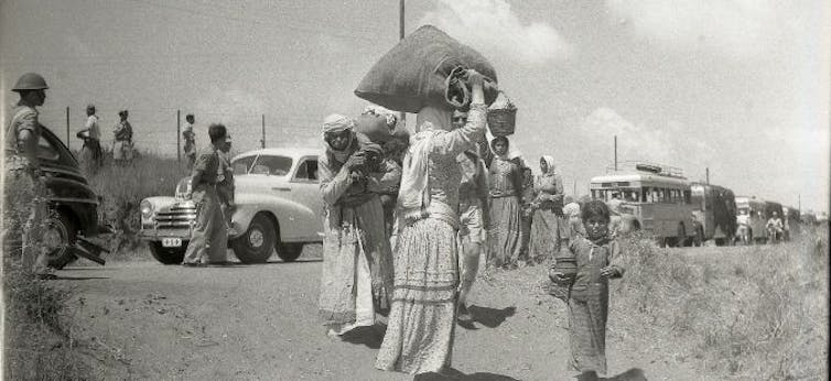 A black and white photo of a group of women walk down a road carrying their belongings.