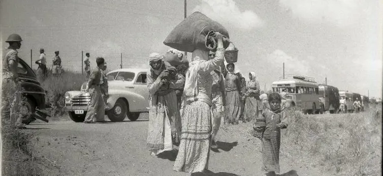 A black and white photo of a group of women walk down a road carrying their belongings.