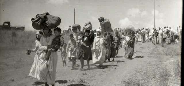 A black and white photo of a group of women walk down a road carrying their belongings.