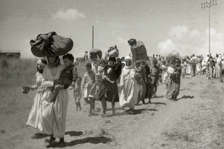 A black and white photo of a group of women walk down a road carrying their belongings.