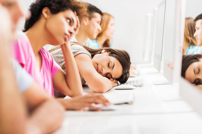 A young female college student rests her head on a computer keyboard while others look at their computer screens.