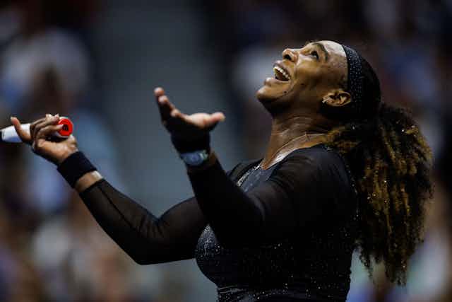 A woman dressed in black with a headband looks heavenward with a big smile on her face, hands raised with palms facing upwards.