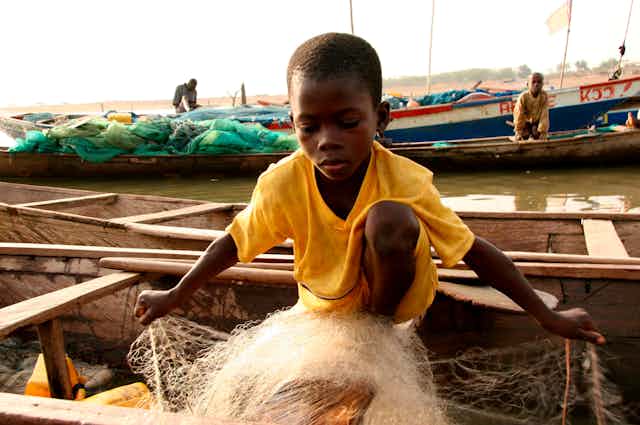 A young Ghanaian boy wearing a yellow top sorting a net on a fishing boat.