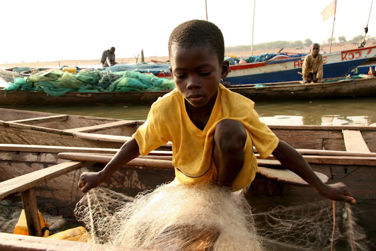 A young Ghanaian boy wearing a yellow top sorting a net on a fishing boat.