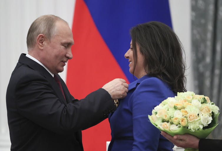 Vlaadimir Putin pins and award to the jacket of the editor-in-chief of broadcaster RT, Margarita Simonyan, with a bouquet of flowers in the foreground.