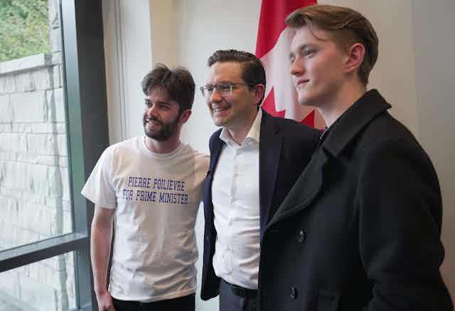 A dark-haired man in a suit jacket wearing glasses is flanked by two young men, one wearing T-shirt that reads Pierre Poilievre for Prime Minister