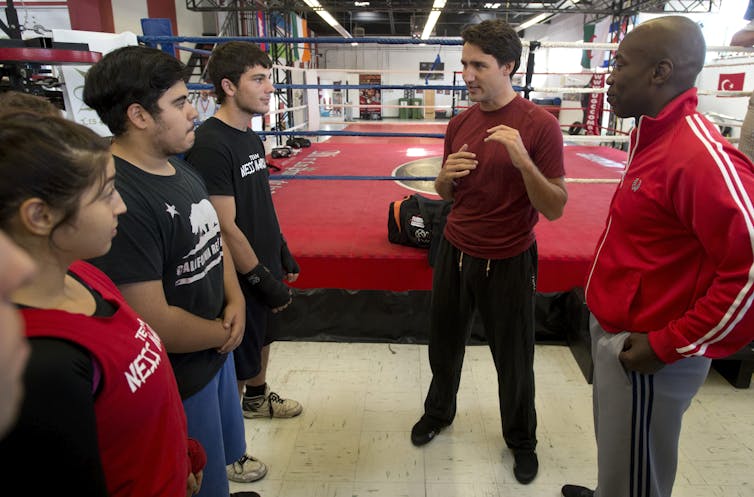 A man in a burgundy T-shirt talks to three young people, a boxing ring behind him and a trainer beside him.