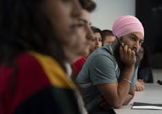 A man in a pink turban sits among a row of young people listening to someone speak off-camera.