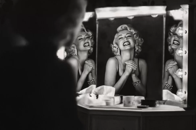 Blonde woman with eyes closed smiling before a mirror.